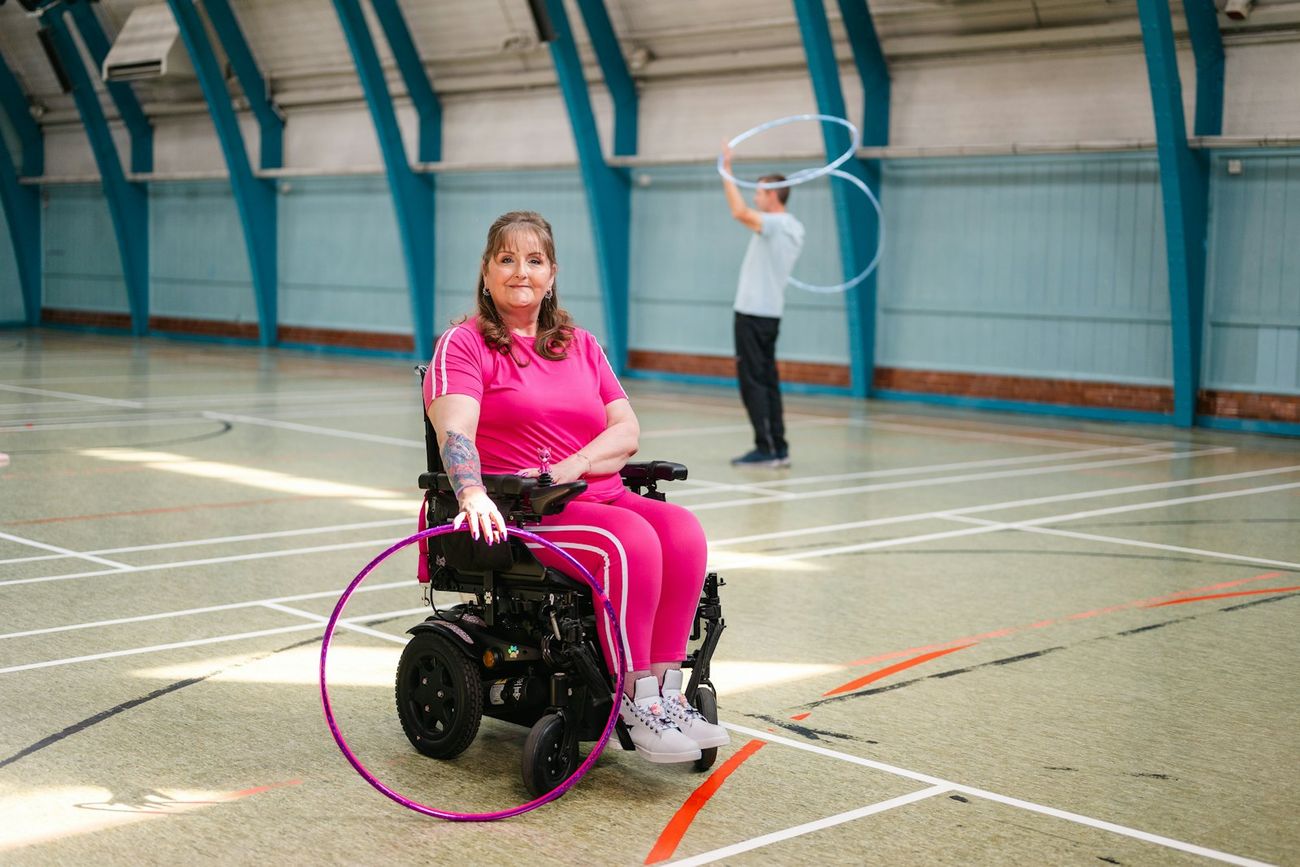A woman in a power wheelchair playing with a hula hoop in a sports hall